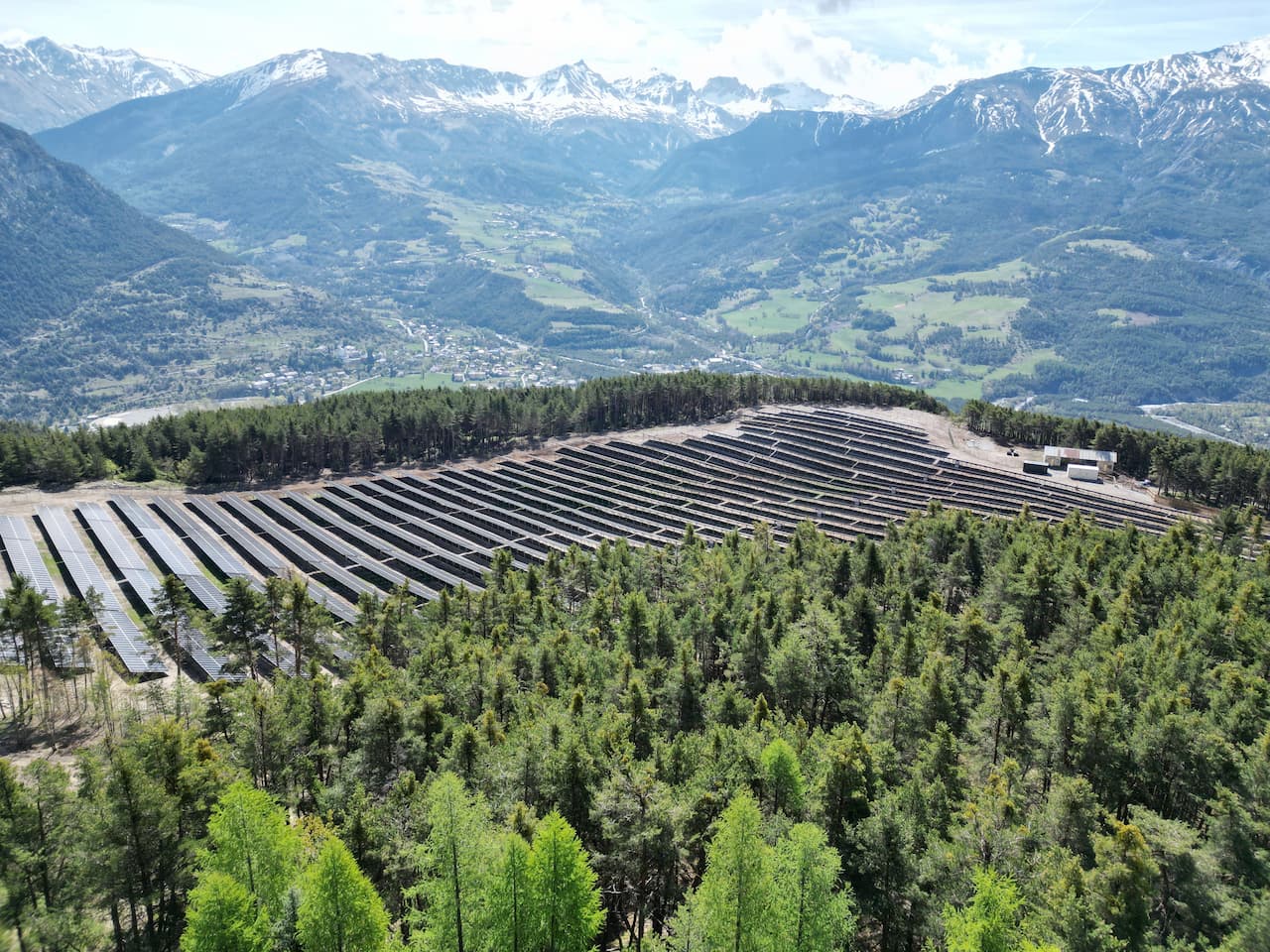 Vue aérienne du parc solaire Le Soleil de Chanenc, rangées de panneaux dans une clairière de forêt face aux montagnes de l'Ubaye